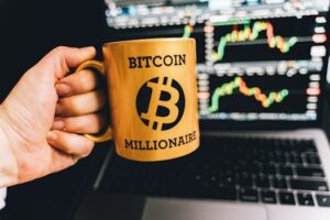 Golden mug with 'Bitcoin Millionaire' text in front of stock market charts on laptop screen.